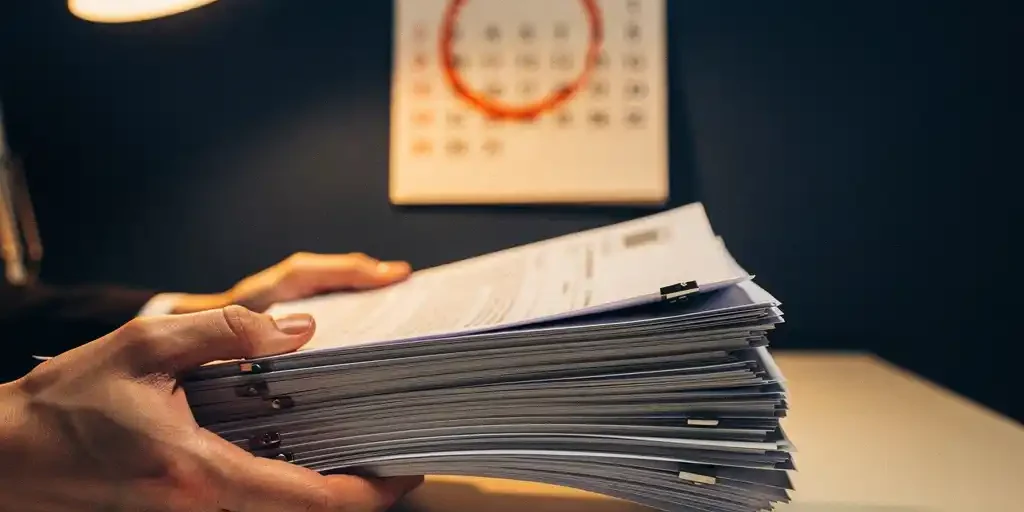 Stapel officiële vergunningsdocumenten in handen boven een bureau, met een kalender met omcirkelde deadline op de achtergrond.